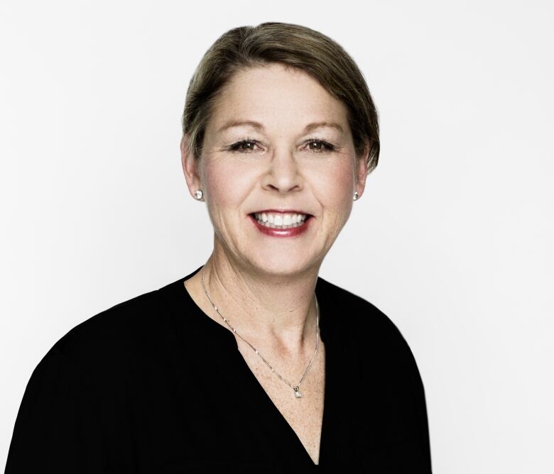 A woman with short brown hair and wearing a black v-neck sweater smiles at the camera for a head and shoulders portrait with a white background