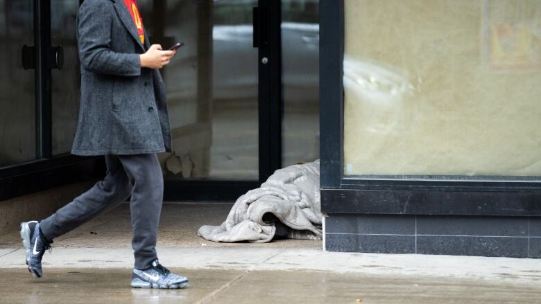 Man on a cell phone walks past a person lying on the sidewalk, wrapped in a blanket