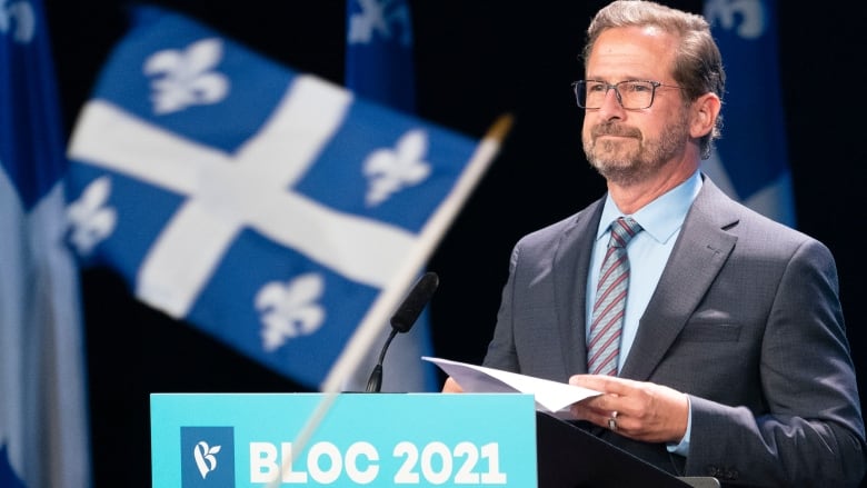 The flag of Quebec flies in the foreground of the Bloc Quebecois leader, standing at attention at a podium with a speech in hand.