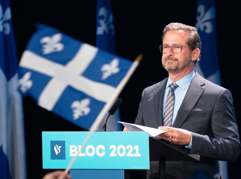 The flag of Quebec flies in the foreground of the Bloc Quebecois leader, standing at attention at a podium with a speech in hand.