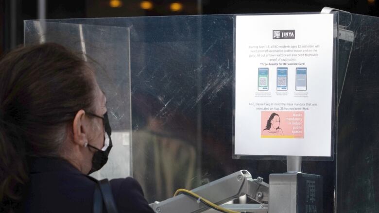 A person in a protective facemask reads a notification about vaccine card and mask requirements outside of a restaurant, mounted on a glass wall.