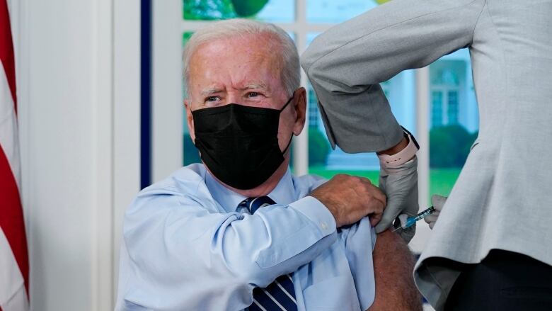 A person wearing a mask rolls up their sleeve as another administers a vaccine shot on their arm.