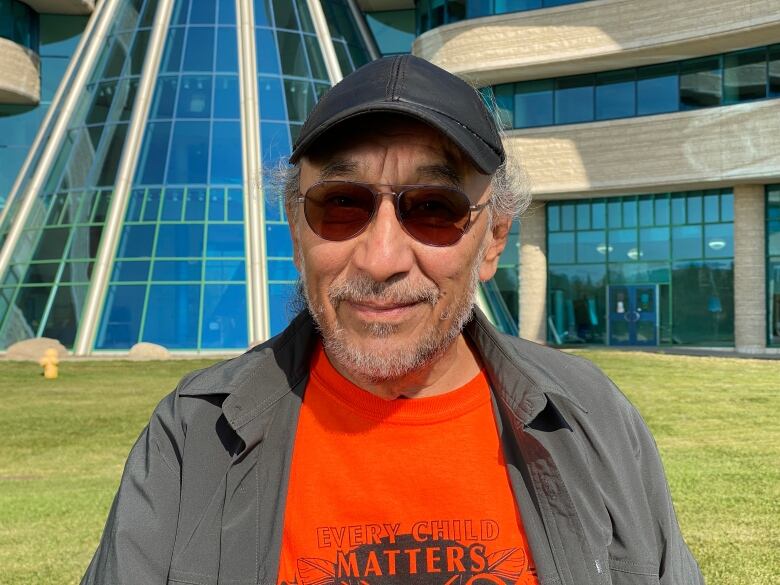 A man with gray hair and sunglasses smiles in front of a glass building