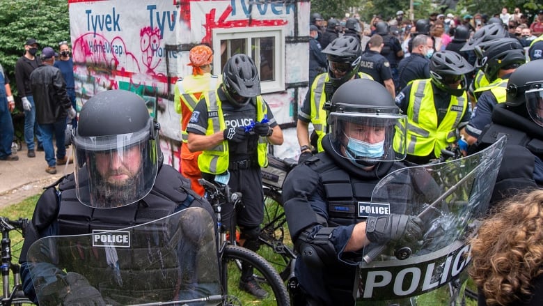 A line of police in riot gear surround a wooden shelter.