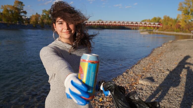 A girl holds up an empty can, while holding a garbage bag in the other hand, in front of the river