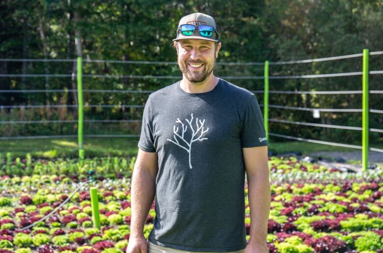 A smiling man wearing a baseball cap stands in a field of lettuce.