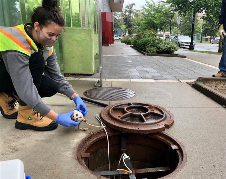 A woman squatting deploys the sewer cage ball to gather a wastewater sample.