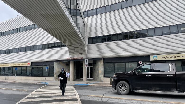 A man walks on a road crosswalk away from a large building and under a pedestrian walkway.