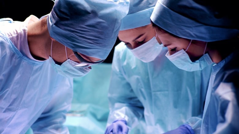 Three surgeons wearing scrubs are pictured looking down at a patient in an operating room.