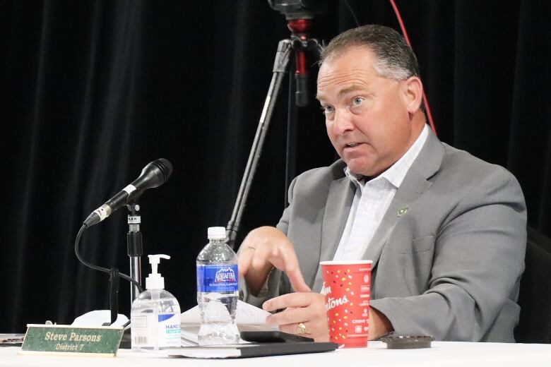 A man in a grey suit and white shirt sits in front of a microphone and gestures with his hands with a disposable coffee cup and plastic water bottle in front of him.