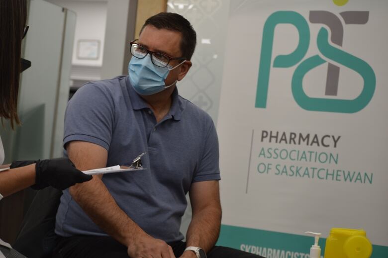 A man in a blue shirt, wearing a disposable mask answers questions from a pharmacist. 