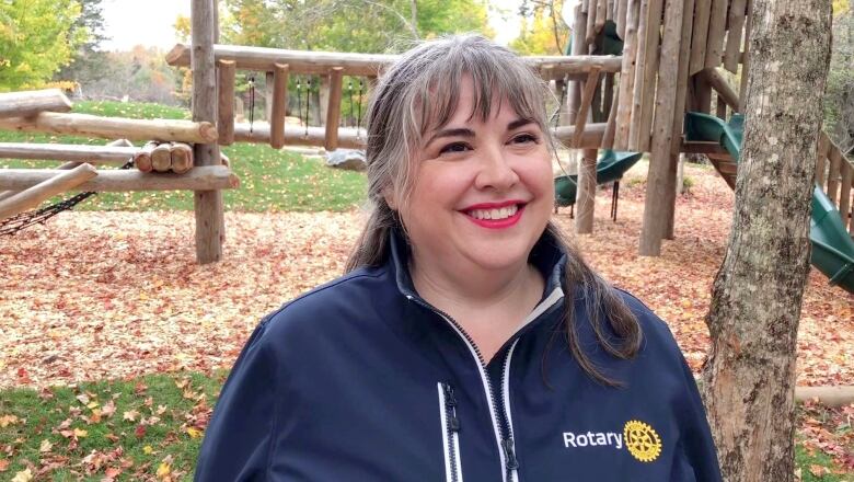 A white woman with long grey hair and a blue zip-up jacket stands outside, with a wooden playset behind her