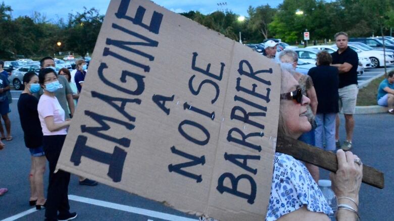 A woman holds a handmade cardboard sign that says Imagine a Noise Barrier. 