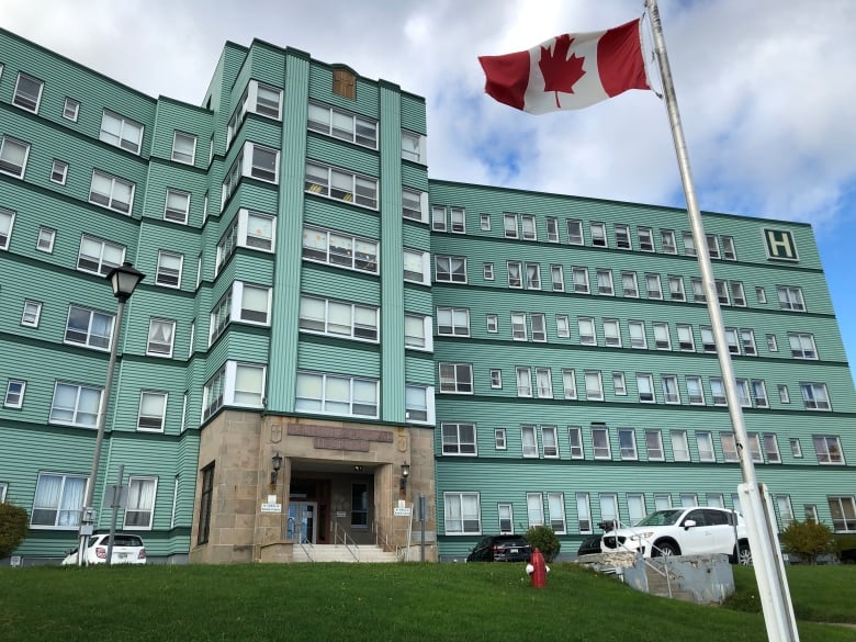 A Canadian flag blows in the wind in front of a green, six-storey hospital building.