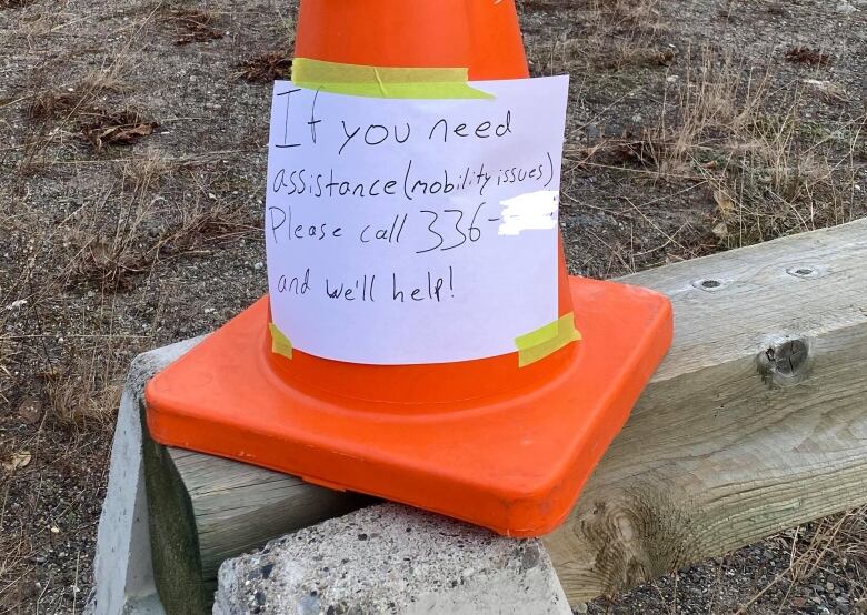 A roadcone with a sign taped to it, which provides a phone number for people who need assistance due to mobility issues. 