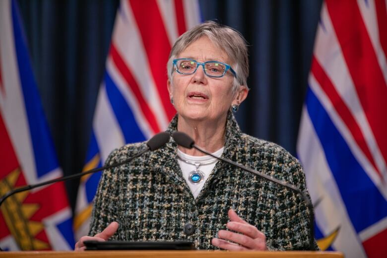 A woman wearing a tweed jacket stands at a podium in front of BC flags