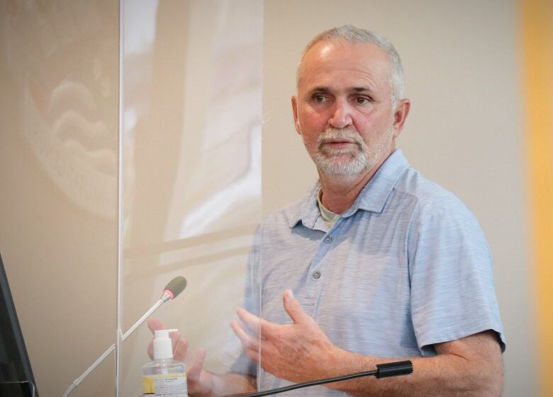 A man with grey hair and a light blue short-sleeved shirt gestures with his hands as he speaks.