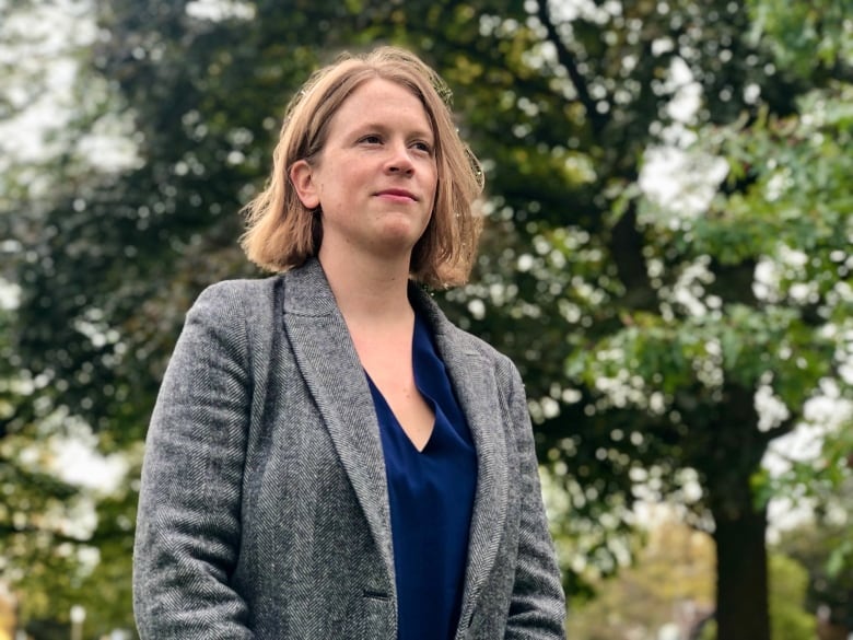 A woman with shoulder-length blone hair, wearing a navy blue shirt and grey blazer, stands outside, with some trees behind her.