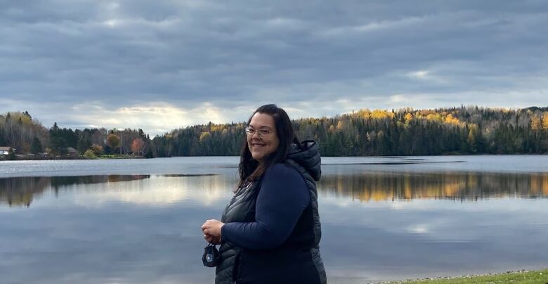 A young woman in winter vest smiles next to a glassy lake rimmed by forest.