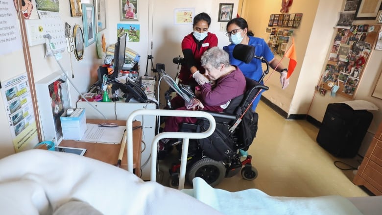 Two women in masks help an older woman in a wheelchair in a nursing home room with photos on the walls.