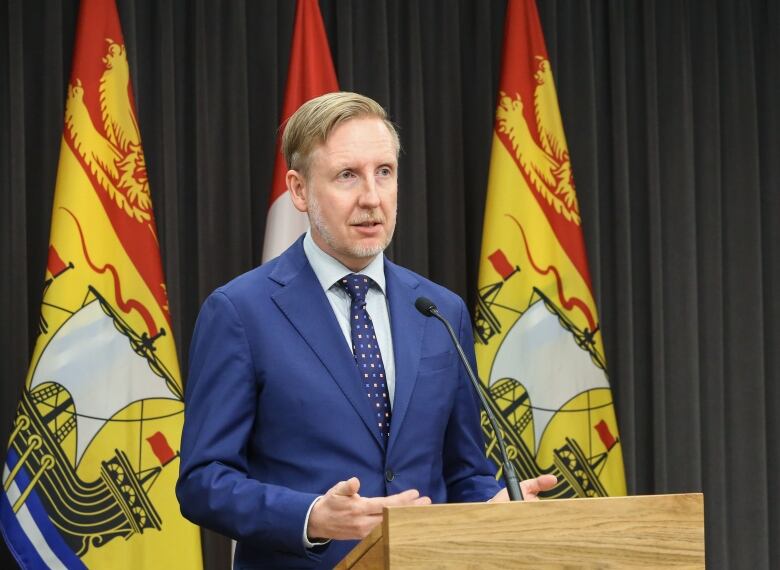 A man wearing a blue suit stands at a lectern. He is flanked by two New Brunswick flags. 