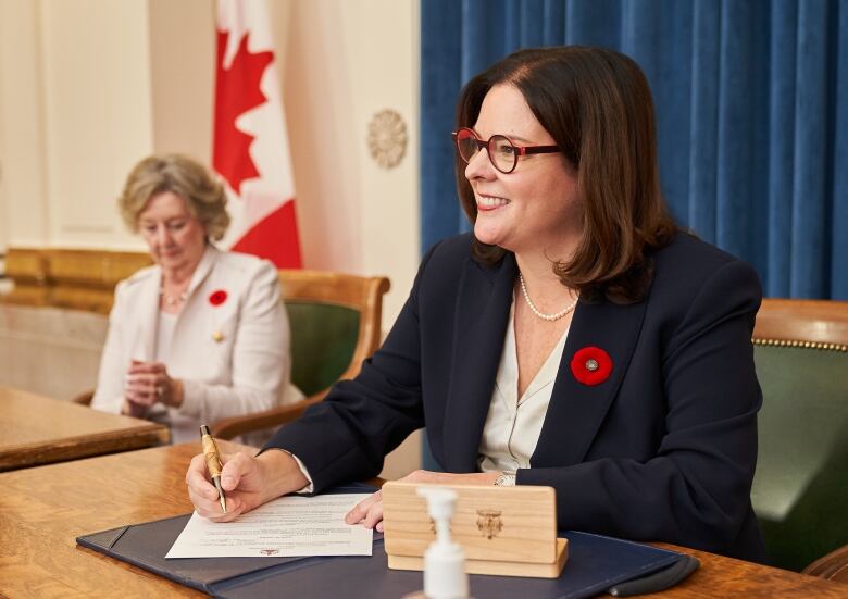 A woman in a dark suit and white blouse smiles while seated as she prepares to sign a document. 