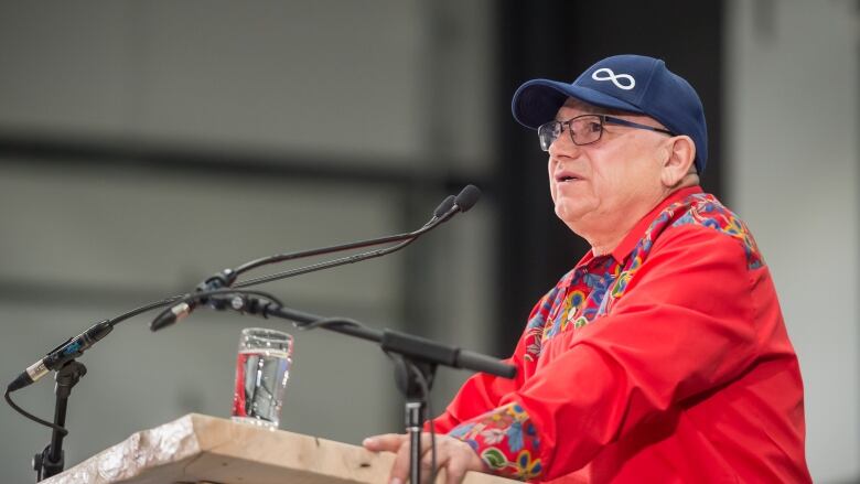 A man in a red shirt and a baseball cap with the Metis insignia speaks at a podium.