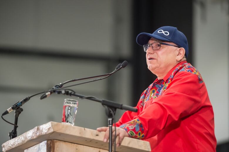 A man in a red shirt and a baseball cap with the Metis insignia speaks at a podium.