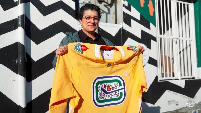 A woman with brown hair tied in a ponytail and red glasses is pictured holding a bright yellow Vancouver Canucks jersey with a colourful Diwali-inspired design.