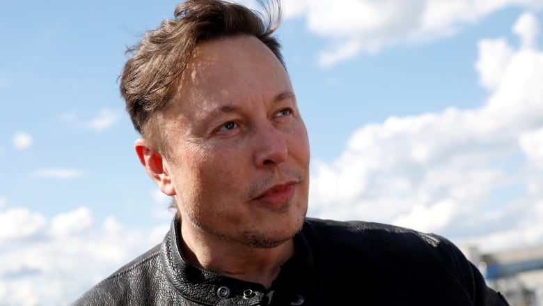 A man in a leather jacket stands outside with a blue sky and white clouds behind him.