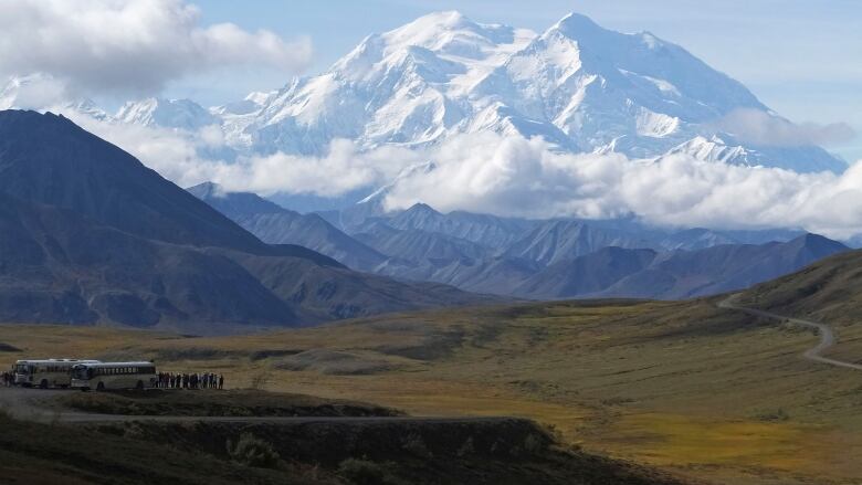 A bus and sightseers look across a vast landscape toward a tall snow-covered mountain.