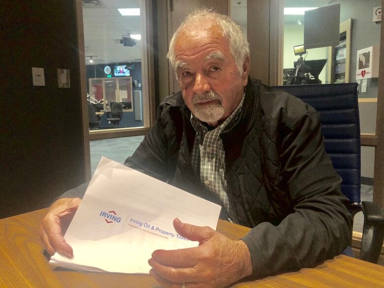 A man with white hair and a beard sits at a desk holding papers.