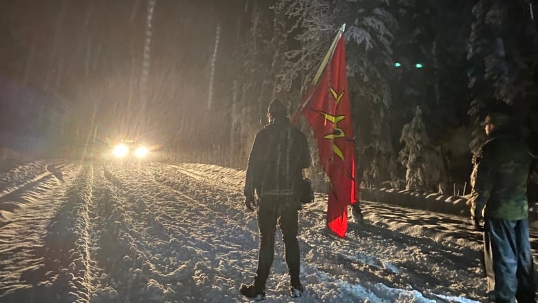 Headlights from a car silhouette a person holding a red flag in the snow.