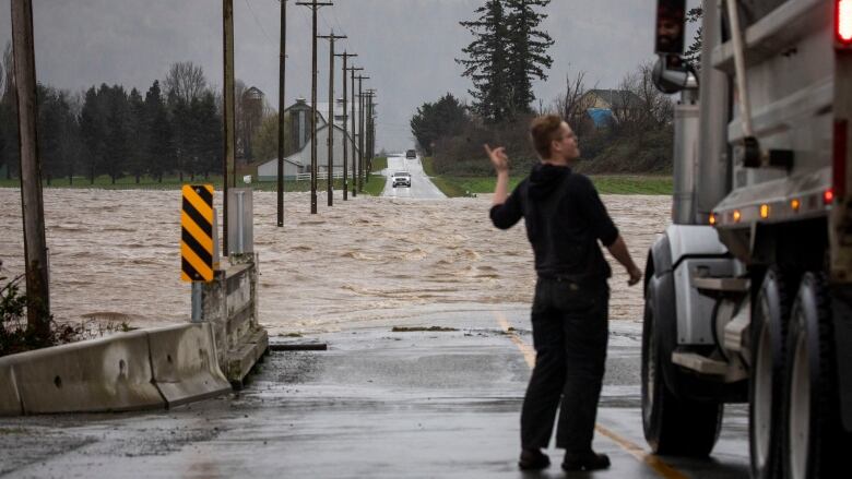 A man stands next to a truck and points at a road that is completely submerged by flood waters.