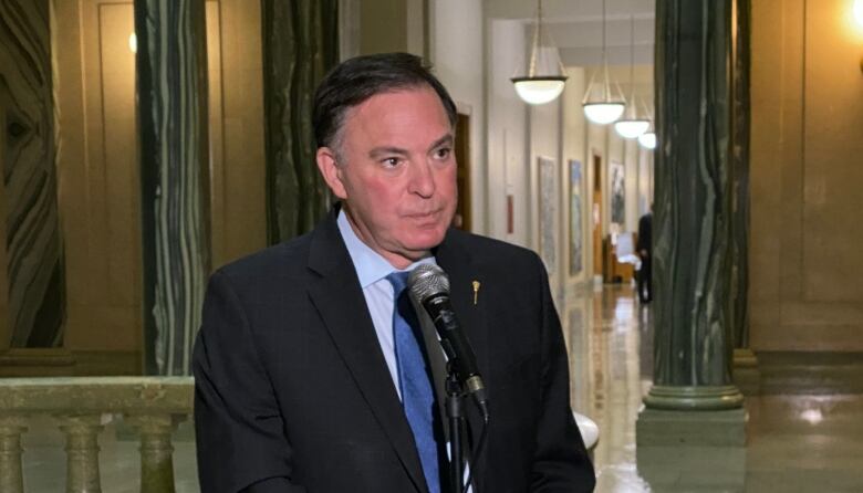 A man wearing a black suit with a pale blue shirt and a blue tie speaks at a microphone. 