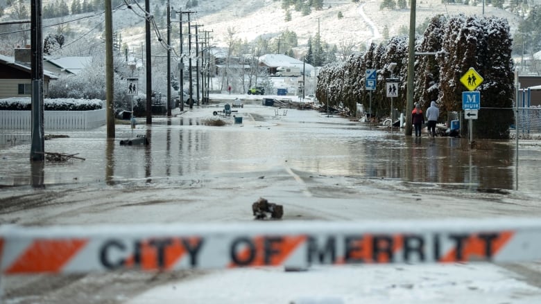 A flooded street is seen behind a barrier, with snow-capped hills visible in the background.