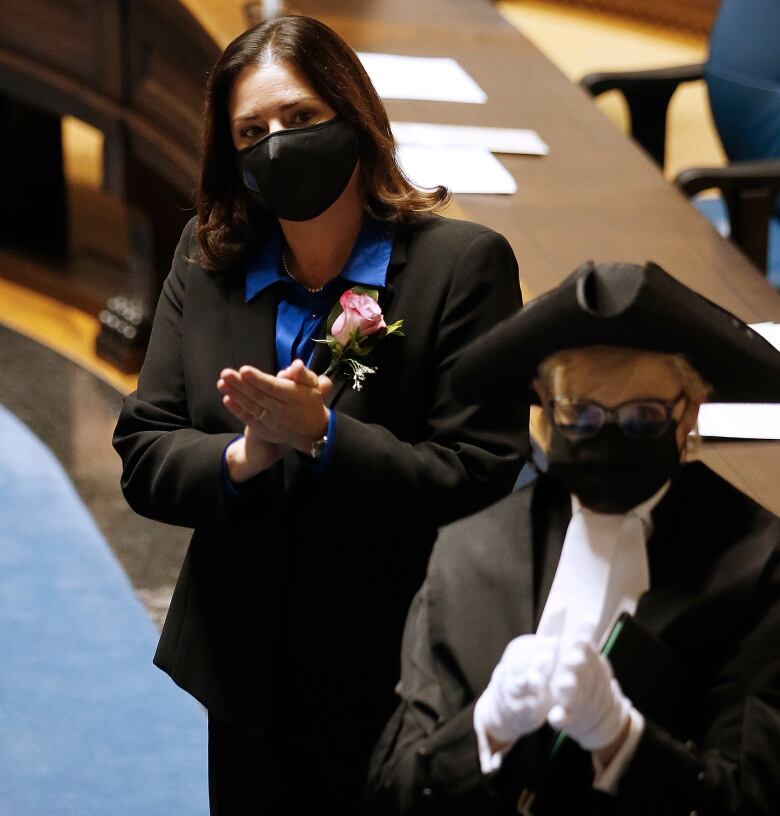 Two women wearing face masks stand and applaud in a legislative chamber.