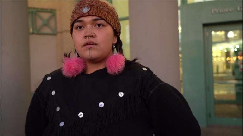 A photo of a woman with pink fur pom-pom earings leaves custody. She has a septum piercing with a smal silver ring and a tradition face tattoo with two lines down her chin. 