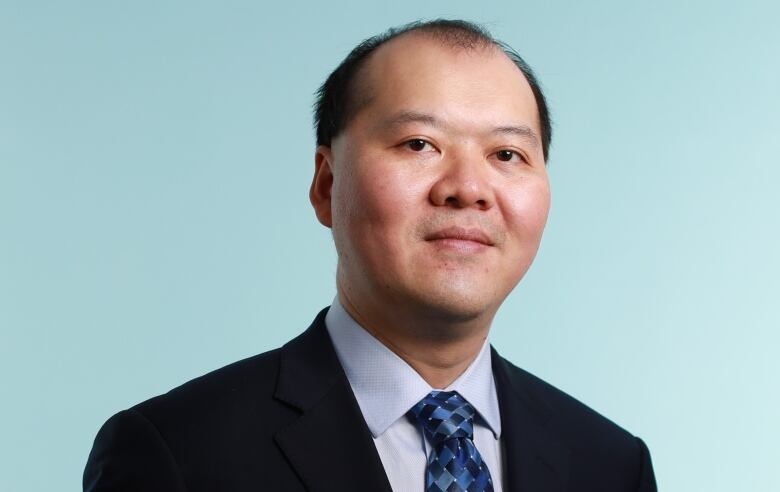 A man in a black suit and blue patterned tie poses for a photo.