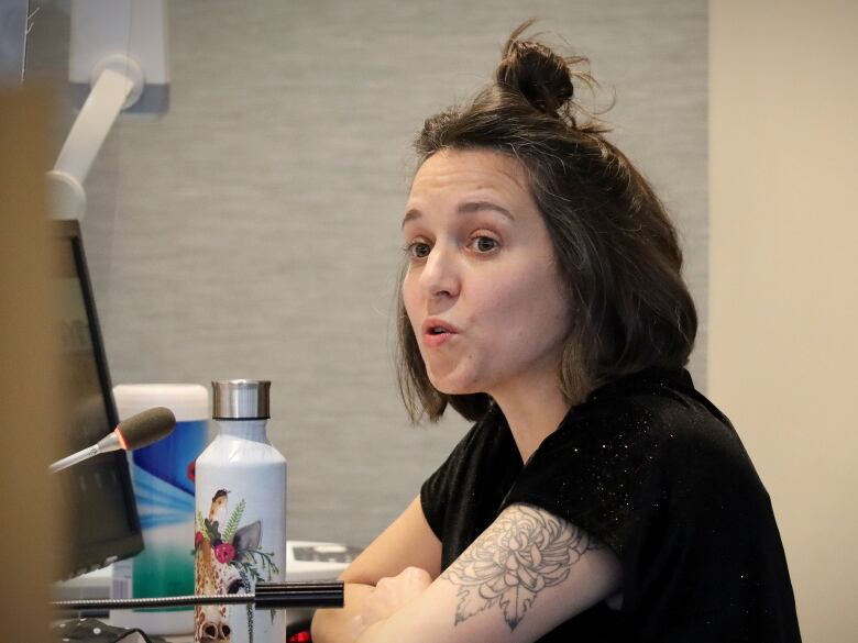 A woman with a black shirt, black tattoo on her arm and a knot of hair on the top of her head speaks into a microphone.