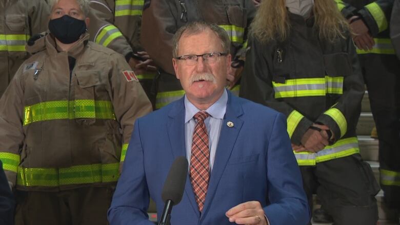 A man in a suit talks into a microphone with firefighters in the background.