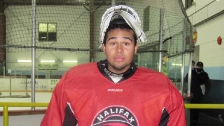 A Black teen is wearing hockey gear with a shirt that reads 'Halifax Hawks