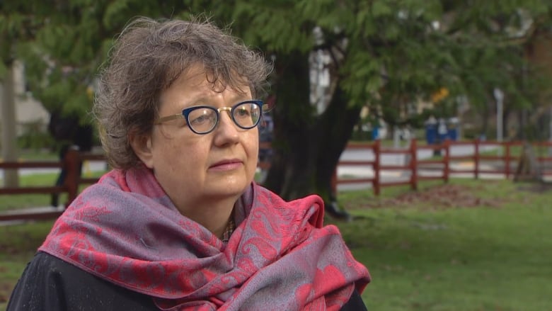 A woman stands in a park wearing glasses and a red and gray shawl.