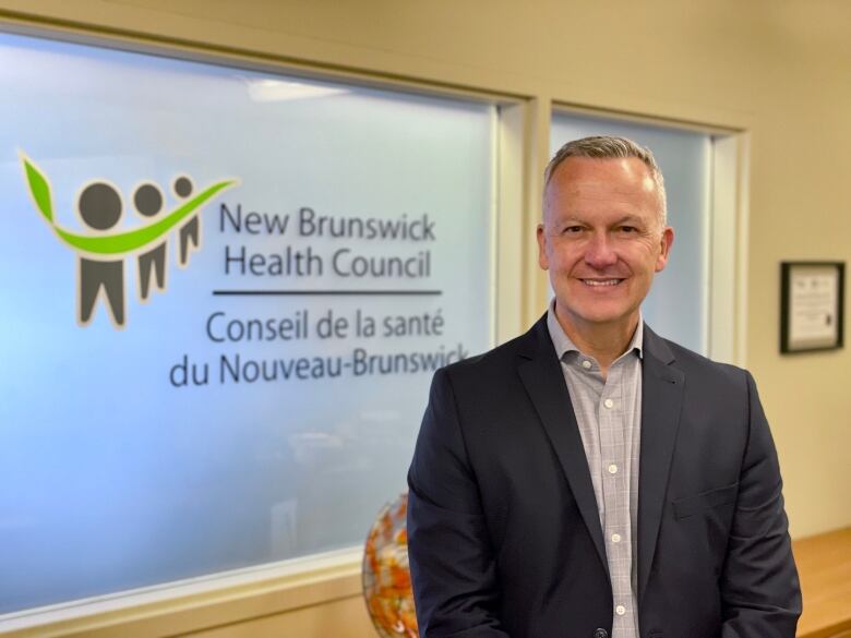 A man with short gray hair, wearing a dark suit and light gray dress shirt smiles at the camera for an upper body portrait standing in front of an opaque window with the words New Brunswick Health Council on it and a logo featuring three humanoid figures and a bright green swish connecting them.