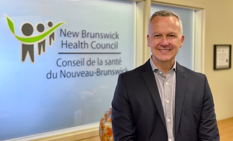 A man with short gray hair, wearing a dark suit and light gray dress shirt smiles at the camera for an upper body portrait standing in front of an opaque window with the words New Brunswick Health Council on it and a logo featuring three humanoid figures and a bright green swish connecting them.