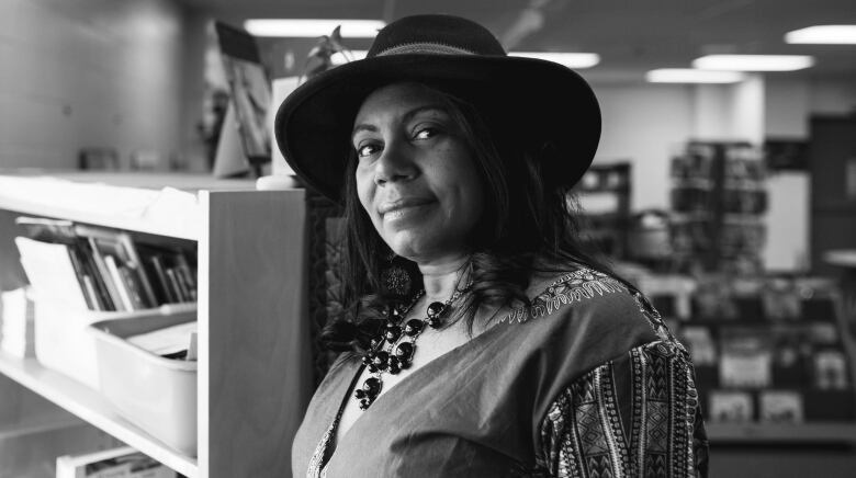 A black and white photo of a woman wearing a dark hat and beaded necklace in what appears to be a library.