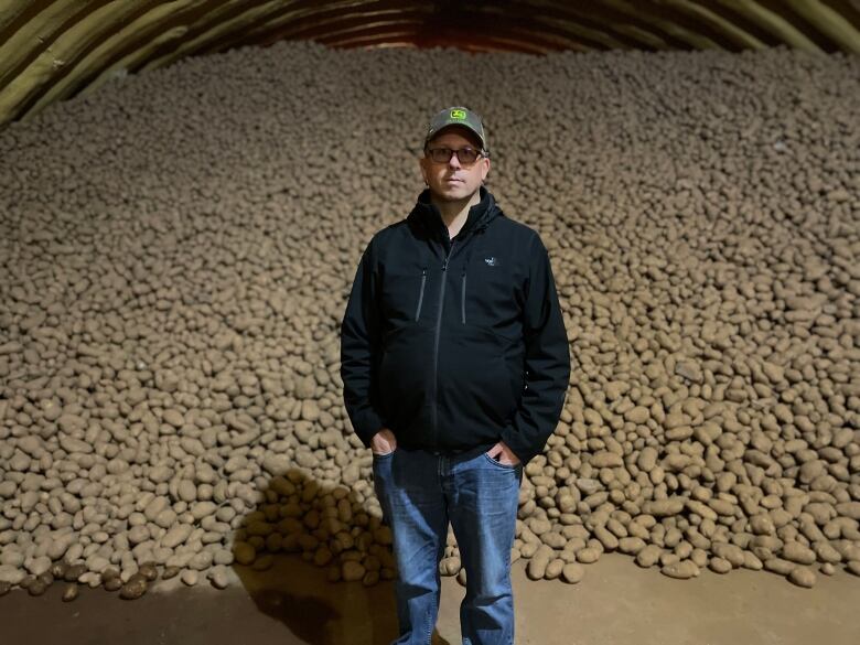 A man in a black windbreaker and green John Deere tractor ballcap stands in a potato warehouse before a mountain of fresh potatoes.