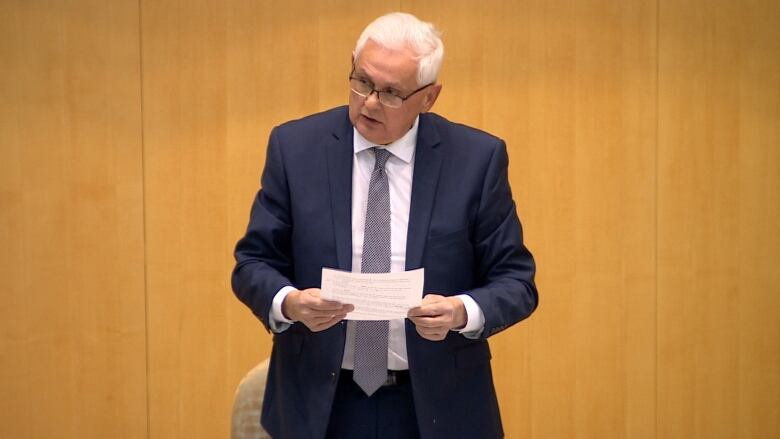 An older man in a suit holds a piece of paper and has glasses on, looking away like he is about to say something.