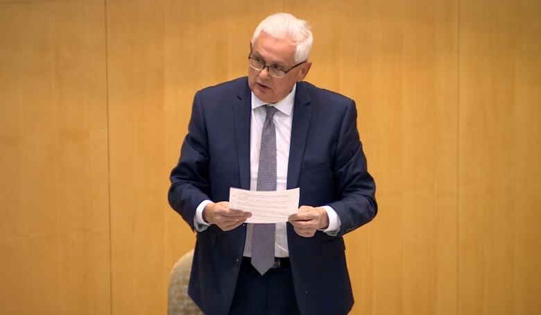 An older man in a suit holds a piece of paper and has glasses on, looking away like he is about to say something.