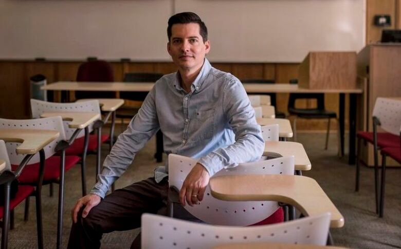 A white man in a blue button up shirt sits backwards in a chair in an empty classroom, with desks surrounding him